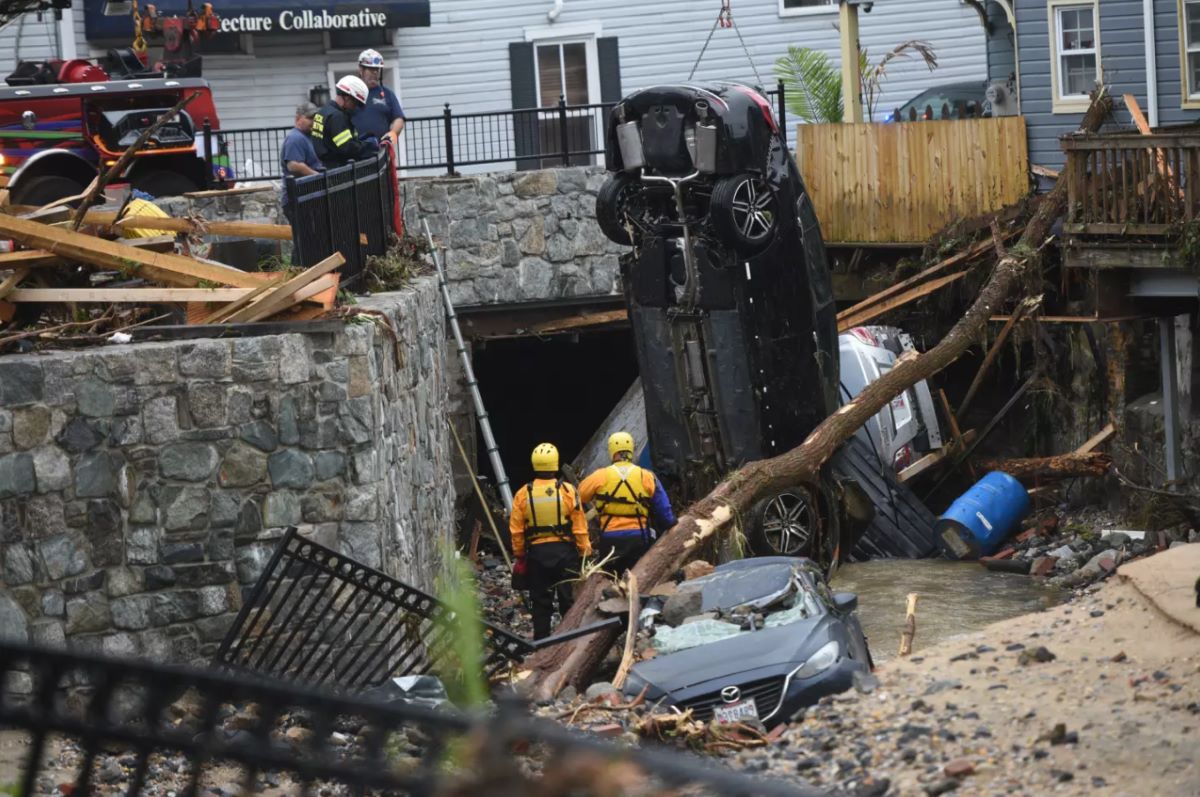 Rescue workers assess the aftermath of severe flooding in Ellicott City, where a car is seen overturned and wedged against a stone wall, partially submerged in debris-filled water. 