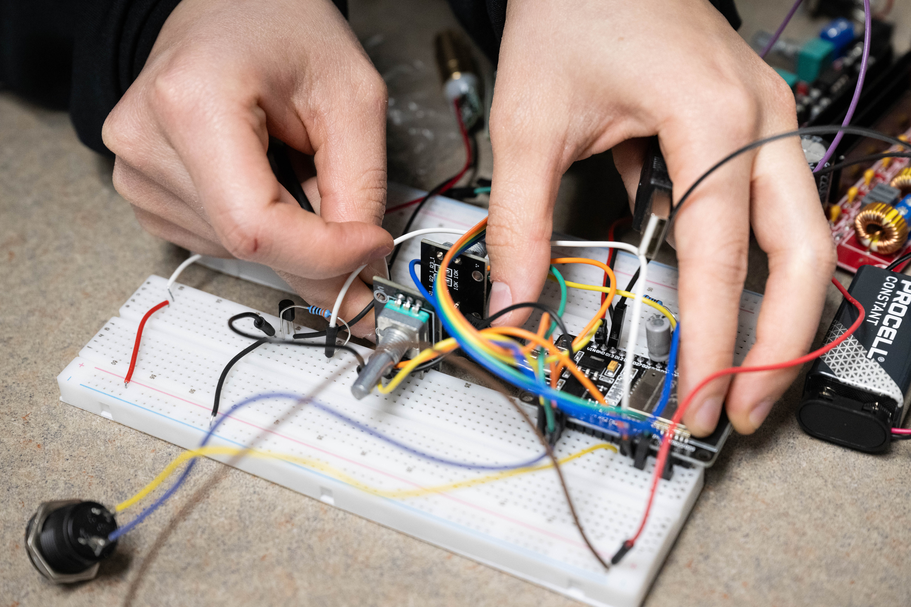A worker puts together electronics by hand, working with a small box and colorful electrical wires