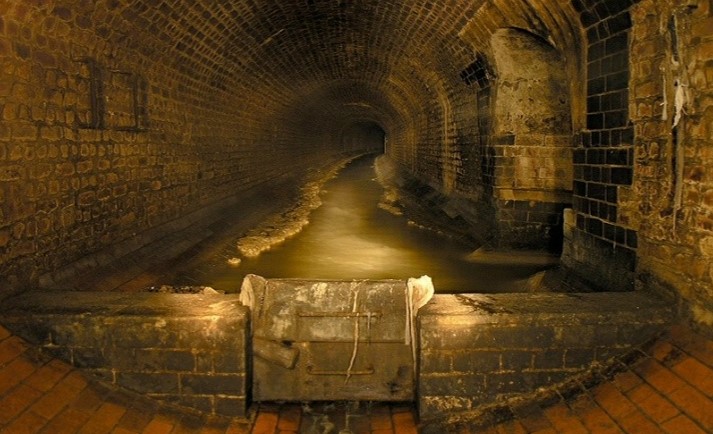 a dimly lit section of the antiquated London sewer system is pictured, with old tile walls lining the long sloping hallway