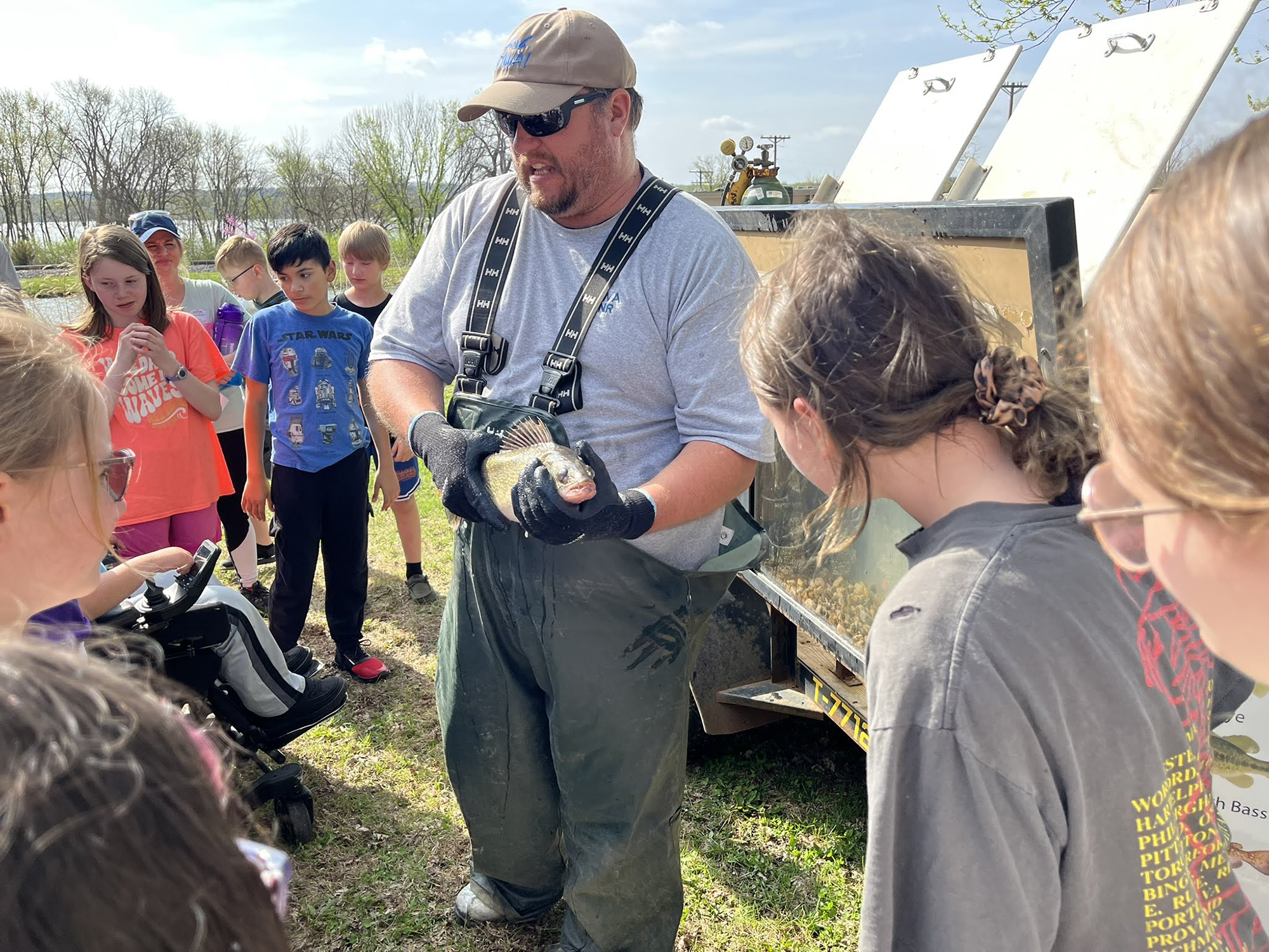 A man in sunglasses and waders holds a fish for students to examine