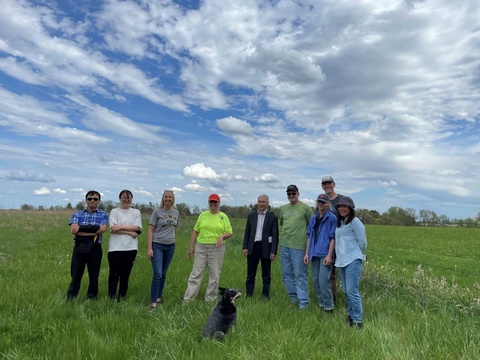 A group of nine people standing together in a grassy field under a partly cloudy sky. A small black and white dog sits in the grass in front of the group.