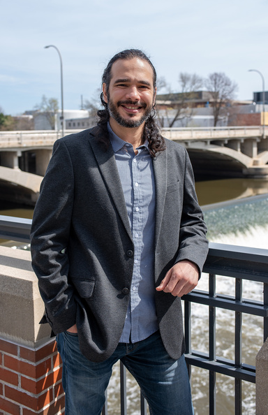 A smiling man leans against a railing with the Iowa River and Burlington Street bridge behind him