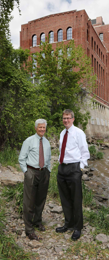 Two men in shirts and ties stand with hands in pockets with a red brick building behind them