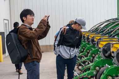A dark haired man with glasses, wearing a brown jacket and a backpack, photographs a green John Deere planter while a friend examines the equipment