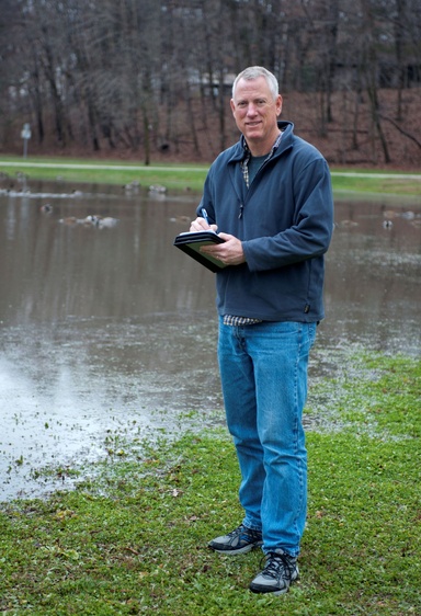 A man in blue jeans stands near a pond holding a clipboard
