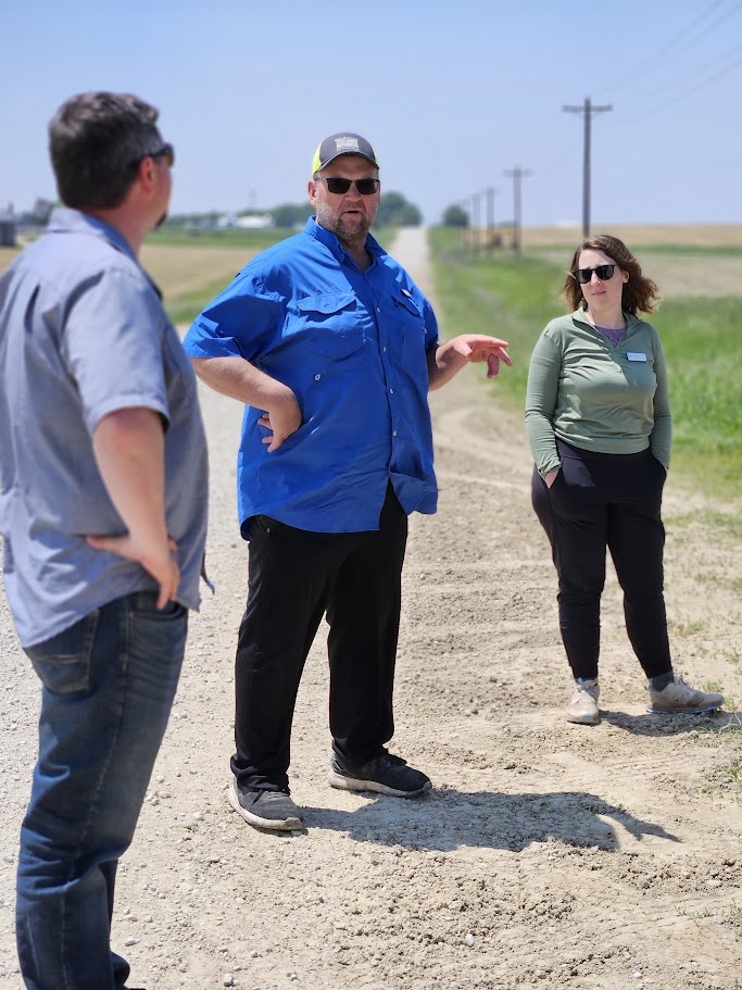 two people stand and listen to a farmer on the side of the road, discussing agricultural practices