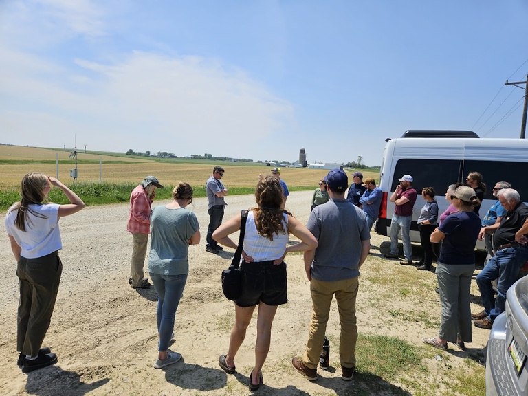A watershed group gathers around in a circle next to their vehicle, discussing agricultural practices