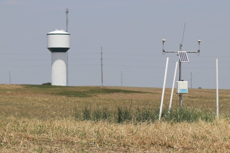 An IFC hydrostation is in focus in a cornfield, with a water tower behind