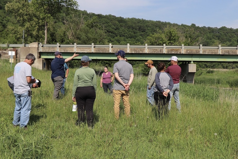 a group gathers in the grass in front of a bridge, with the IFC's river monitoring system on it