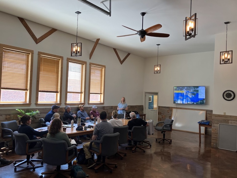 a group sits around a large wooden table in a conference room, looking at a presentation