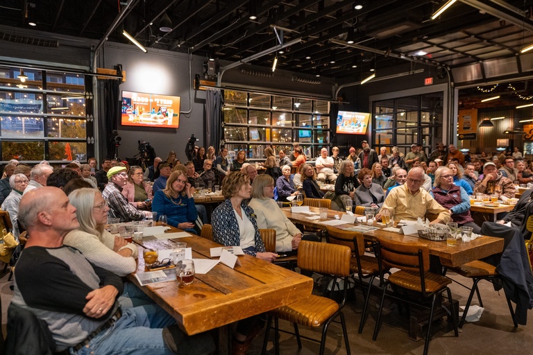 A large crowd sits at long tables in Big Grove Brewery, facing the speakers