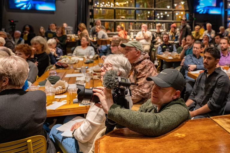 A photographer takes a photo of the panel, bent down by a table in the crowd