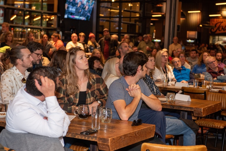 A large crowd sits at long tables in Big Grove Brewery, facing the speakers