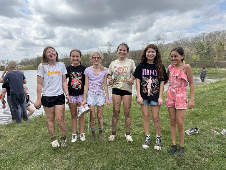 Six smiling girls covered in mud stand on the banks of the pond