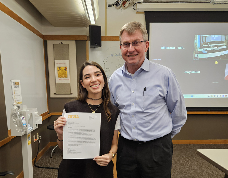 A smiling young woman holds an award letter; beside her stands a tall smiling man