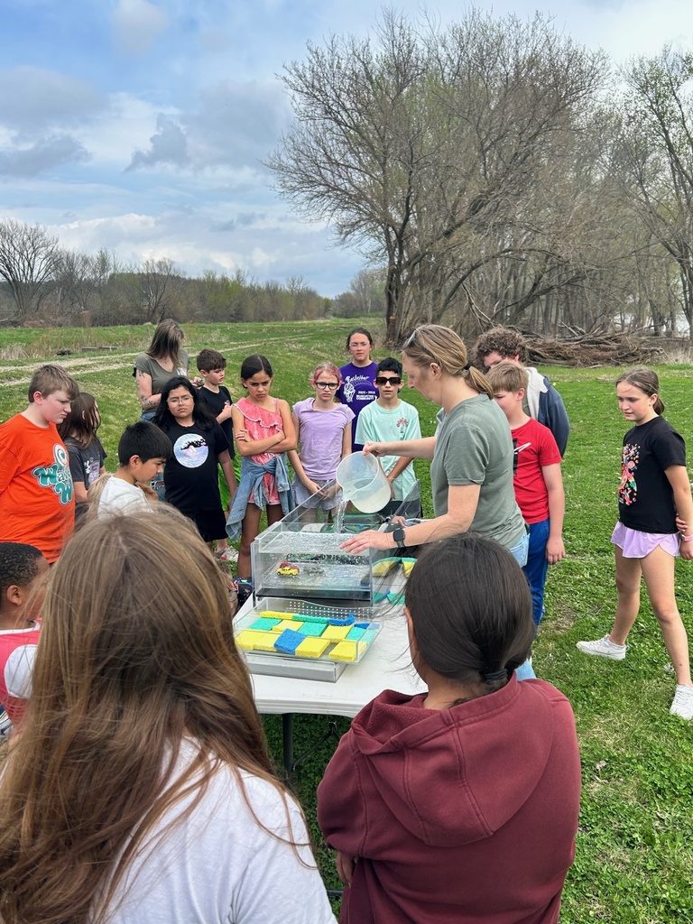 Kids gather around a tabletop watershed model as a woman pours water into it.