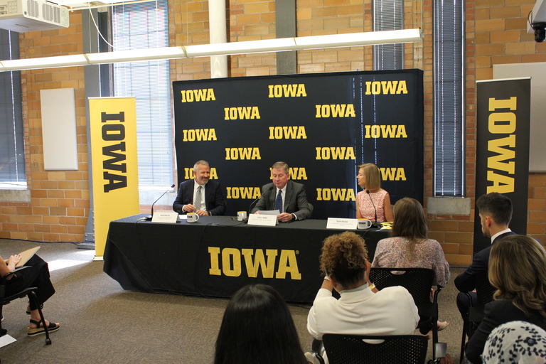 Larry Weber, David Cwiertny, and Sharon Krause sit at a table at the press event, delivering remarks in front of an Iowa banner