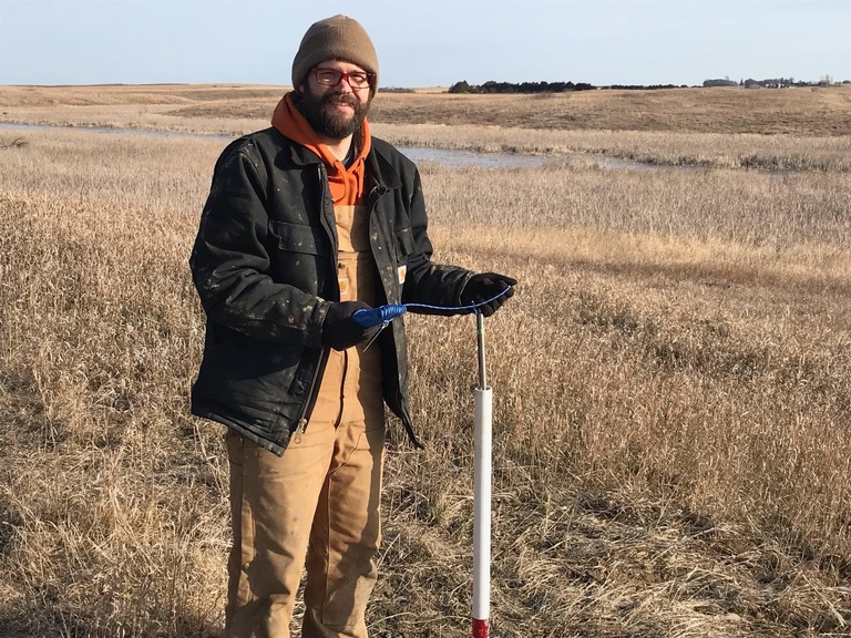 A man in winter clothes works with a monitoring well in a brown grassy field