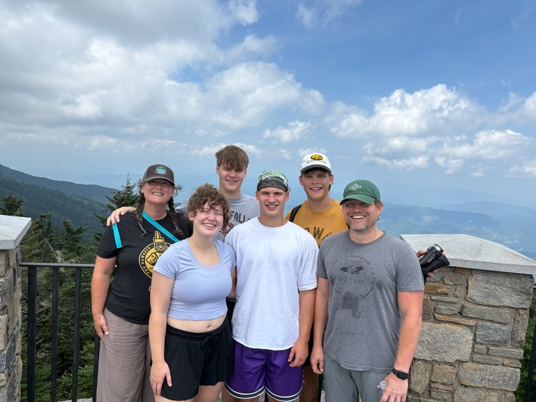 A group of people stands in front of a beautiful mountain vista with blue skies and white clouds behind them