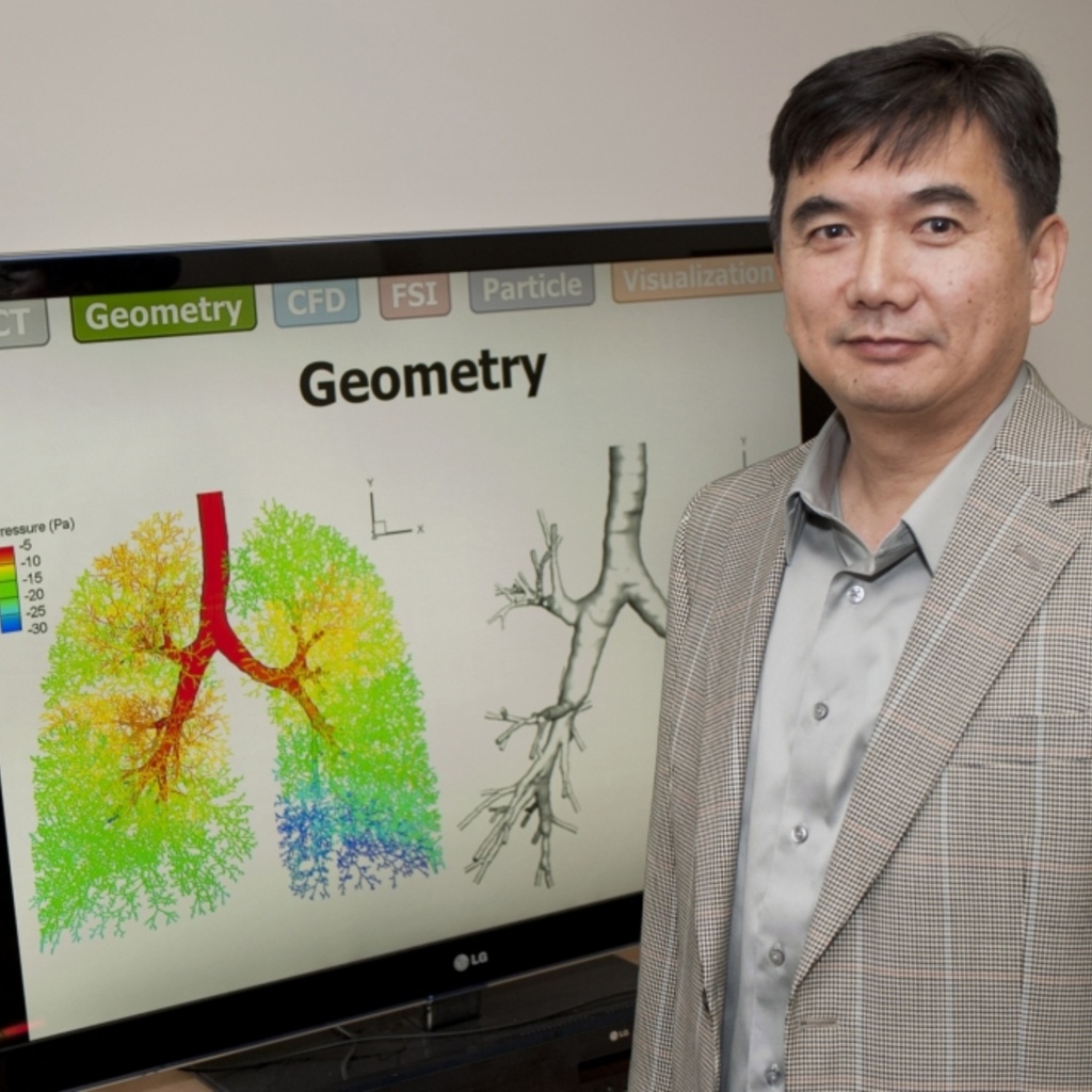 Man in a suit coat stands beside an electronic display of a colorful simulation of airflow through the human lung