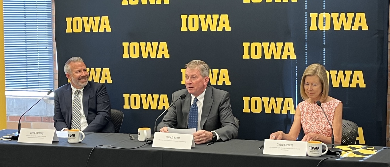 Two men and a woman sit at a table with a black and gold IOWA banner behind them