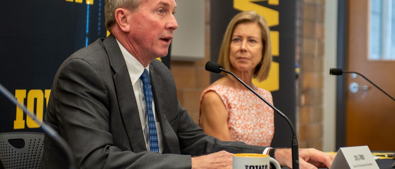 A man in a gray suit speaks while a woman looks on. Both sit in front of a black and gold IOWA banner