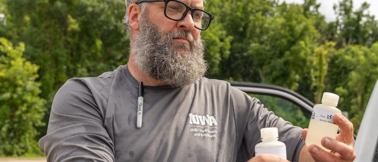 A researcher examines water quality samples with green trees behind him