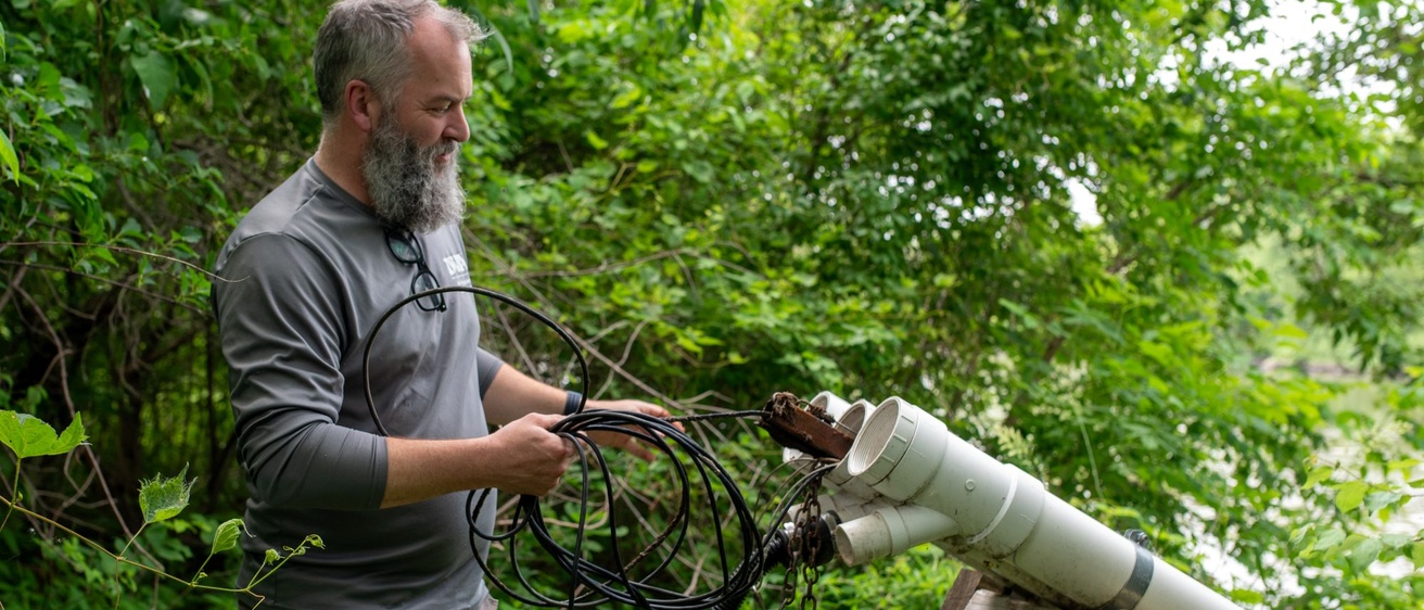 A man makes adjustments to a water quality sensor in white PVC pipe. They're in a green wooded area.