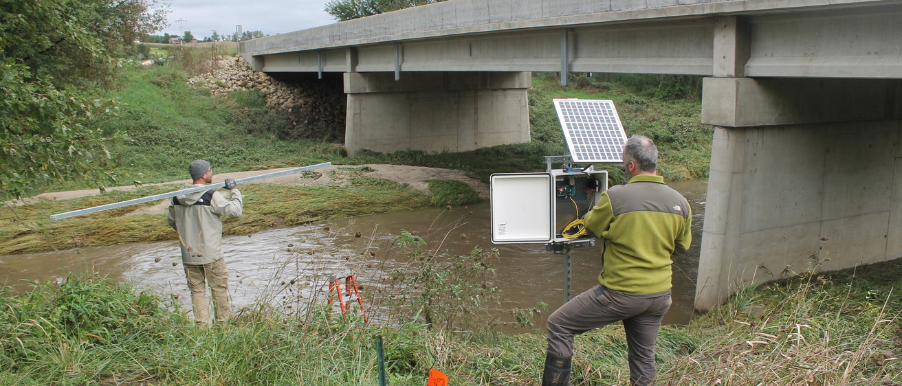 Two men install water quality sensor equipment on the banks of a stream with a bridge in the background