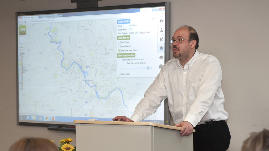 A man stands behind a podium, lecturing to a classroom of students.