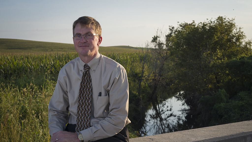 A man in shirt and tie sits on a bridge rail on a rural country road.