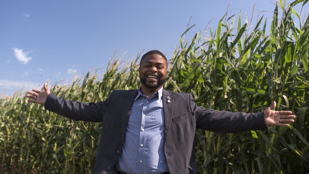 A smiling man stands in front of a field of corn in late summer with arms outspread