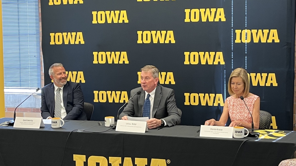 Two men and a woman sit at a table with a black and gold IOWA banner behind them