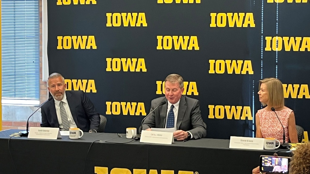 Two men and a woman sit at a table with a black and gold Iowa banner behind them