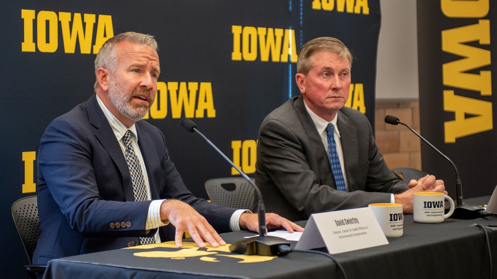 Two men in suits and ties sit at a table, answering questions from reporters. Behind them is a black and gold University of Iowa backdrop